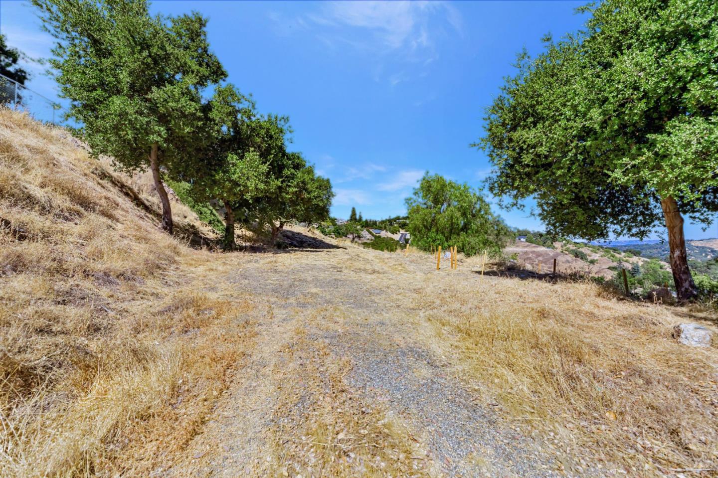 Vacant hillside lot with a gravel track, dry grass, mature trees, and distant hills.
