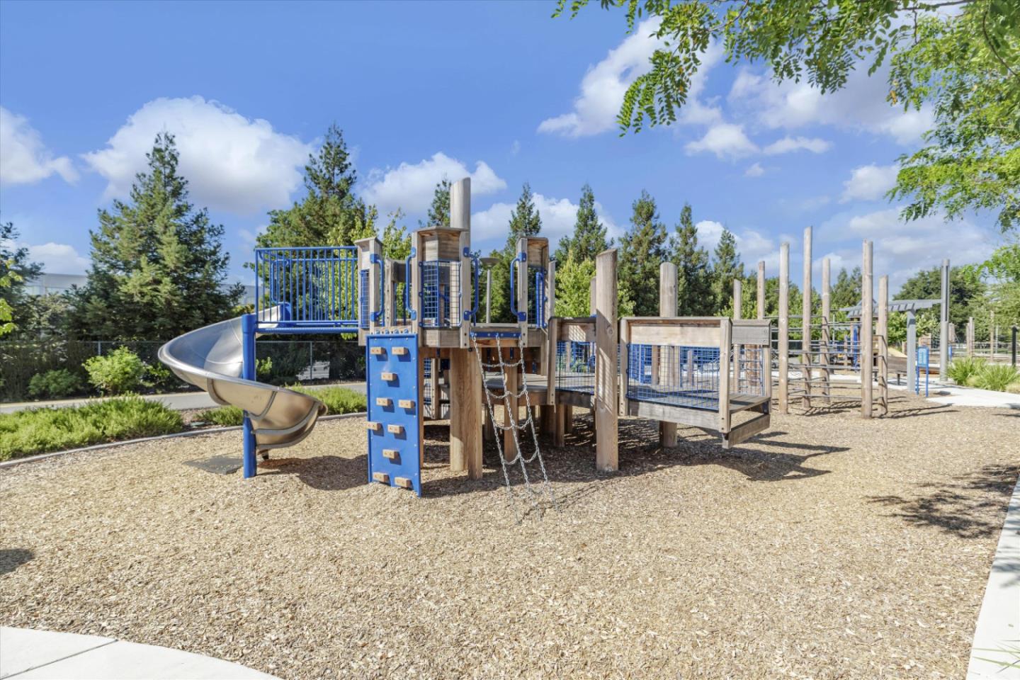 Community playground with a wooden climbing structure, blue climbing wall, and curved metal slide.