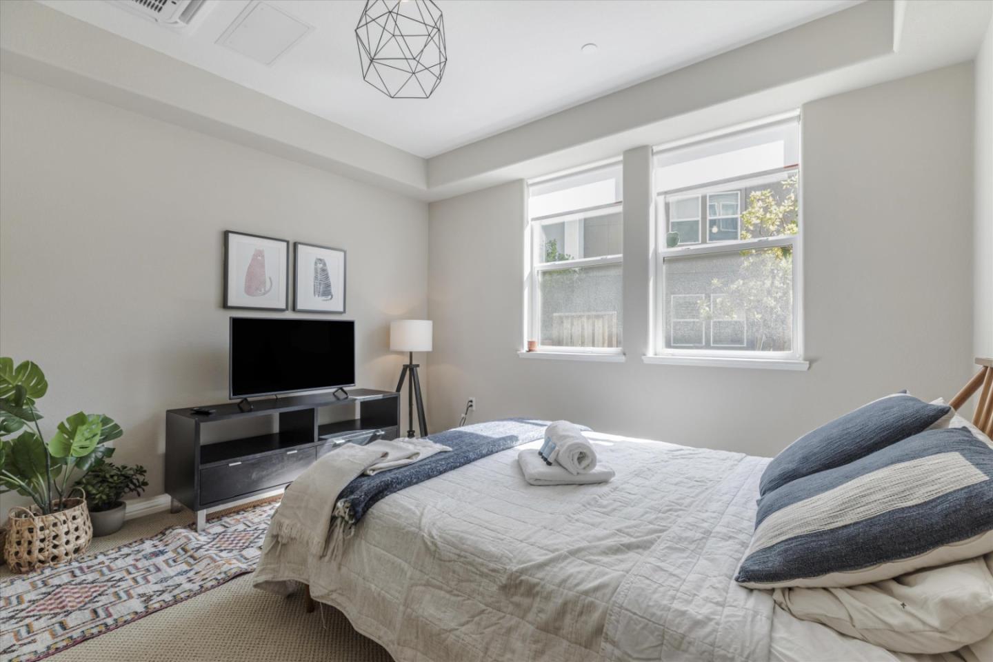 Staged bedroom with a neatly made bed, blue pillows, and two large windows.