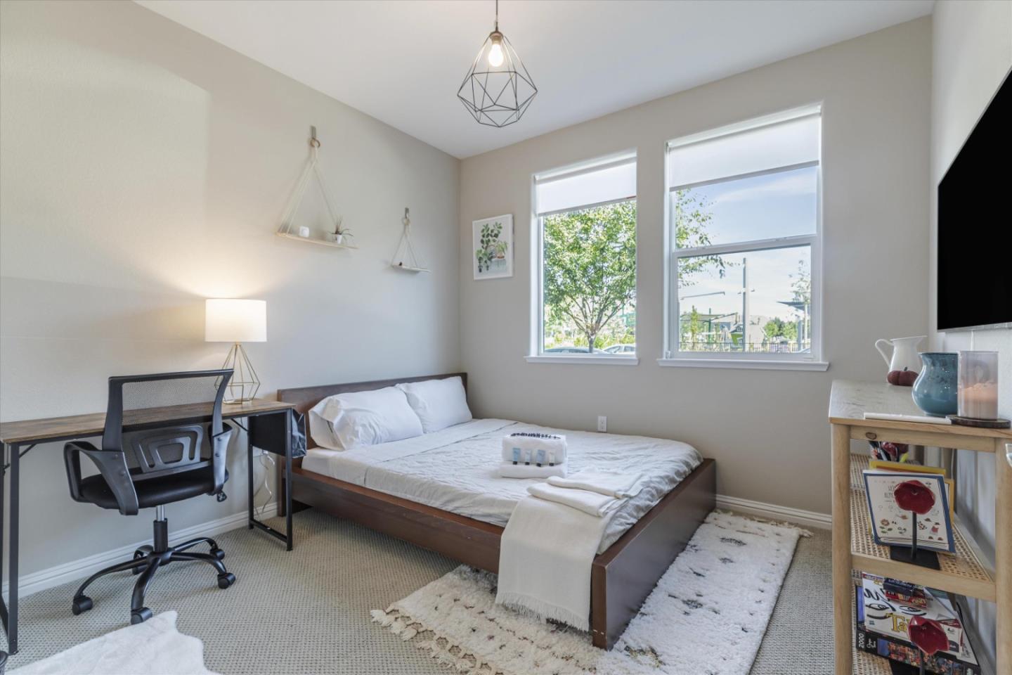 Bedroom with a low white-dressed bed, desk, and two tall windows overlooking trees.