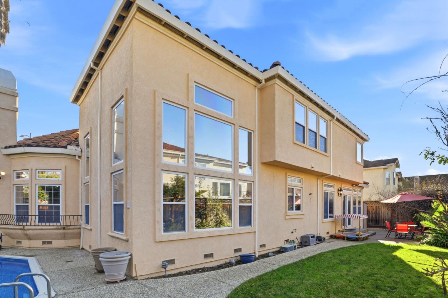 Two-story beige stucco home with large windows, patio seating, lawn, and a pool edge.