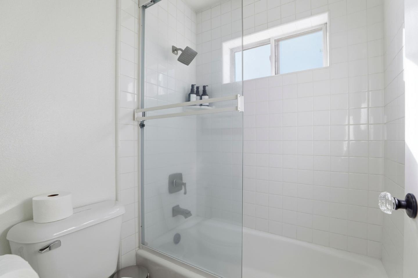 White-tiled bathroom with a glass shower enclosure, bathtub, and high window.