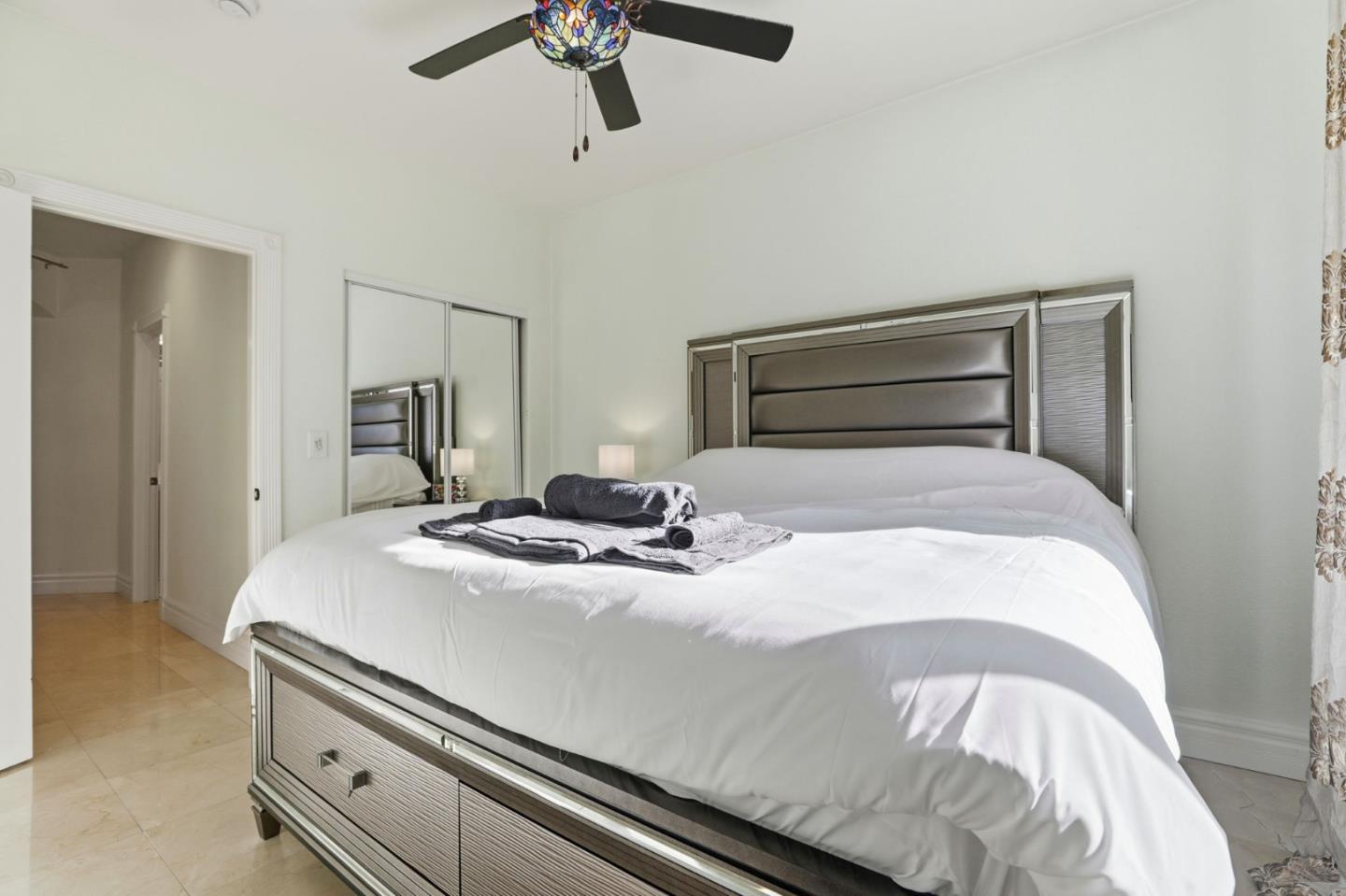 Bedroom with a large white bed, metallic paneled headboard, mirrored closet doors, and decorative ceiling fan.