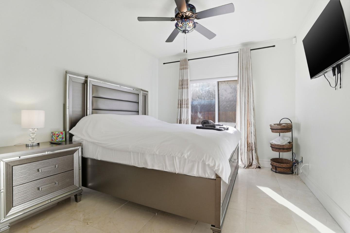 Bedroom with a large bed, decorative ceiling fan, curtained window, and wall-mounted television.