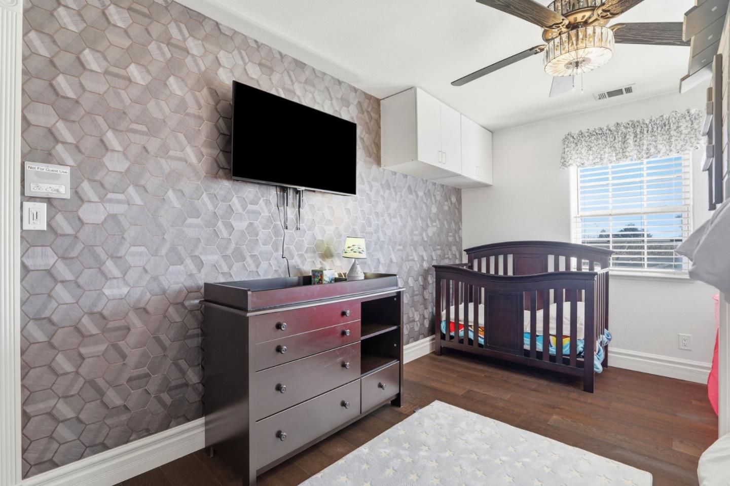 Nursery with a dark wood crib, matching dresser, geometric wallpaper, and wall-mounted television.