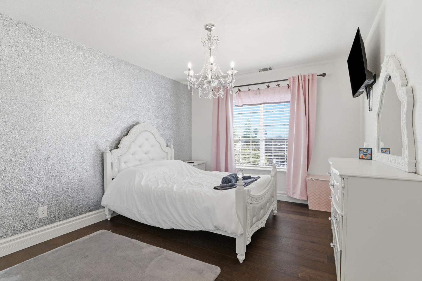 Bedroom with an ornate white bed, silver accent wall, pink curtains, and chandelier.