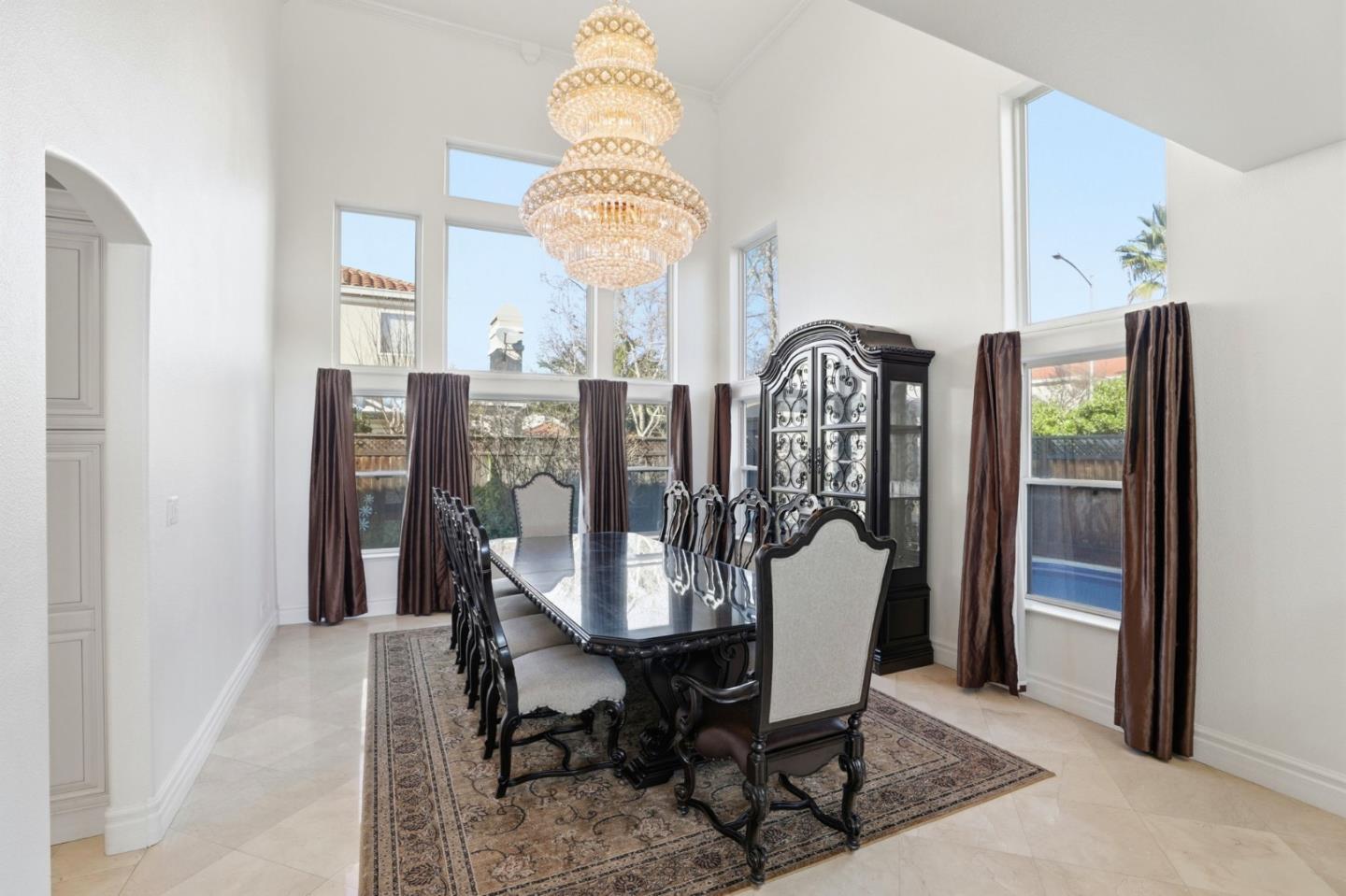 Dining room with a crystal chandelier, carved dark furniture, and tall curtained windows.