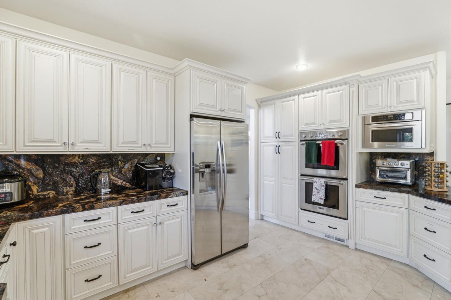 Kitchen with white paneled cabinets, dark marbled granite counters, stainless refrigerator, and double ovens.