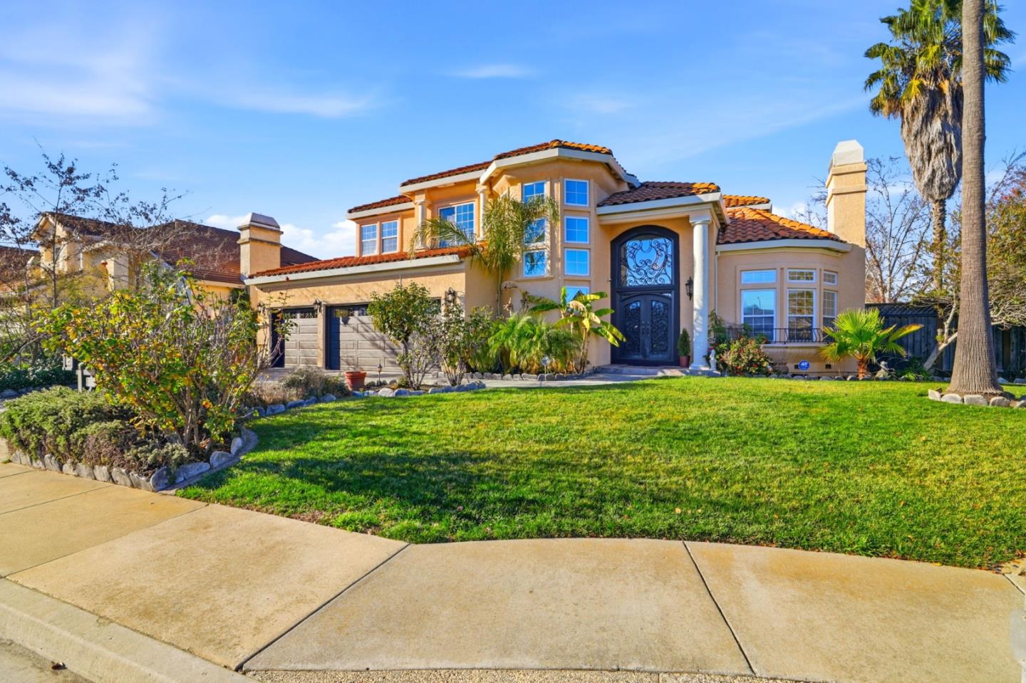 Two-story tan stucco home with a red tile roof, ornate entry door, and broad front lawn.
