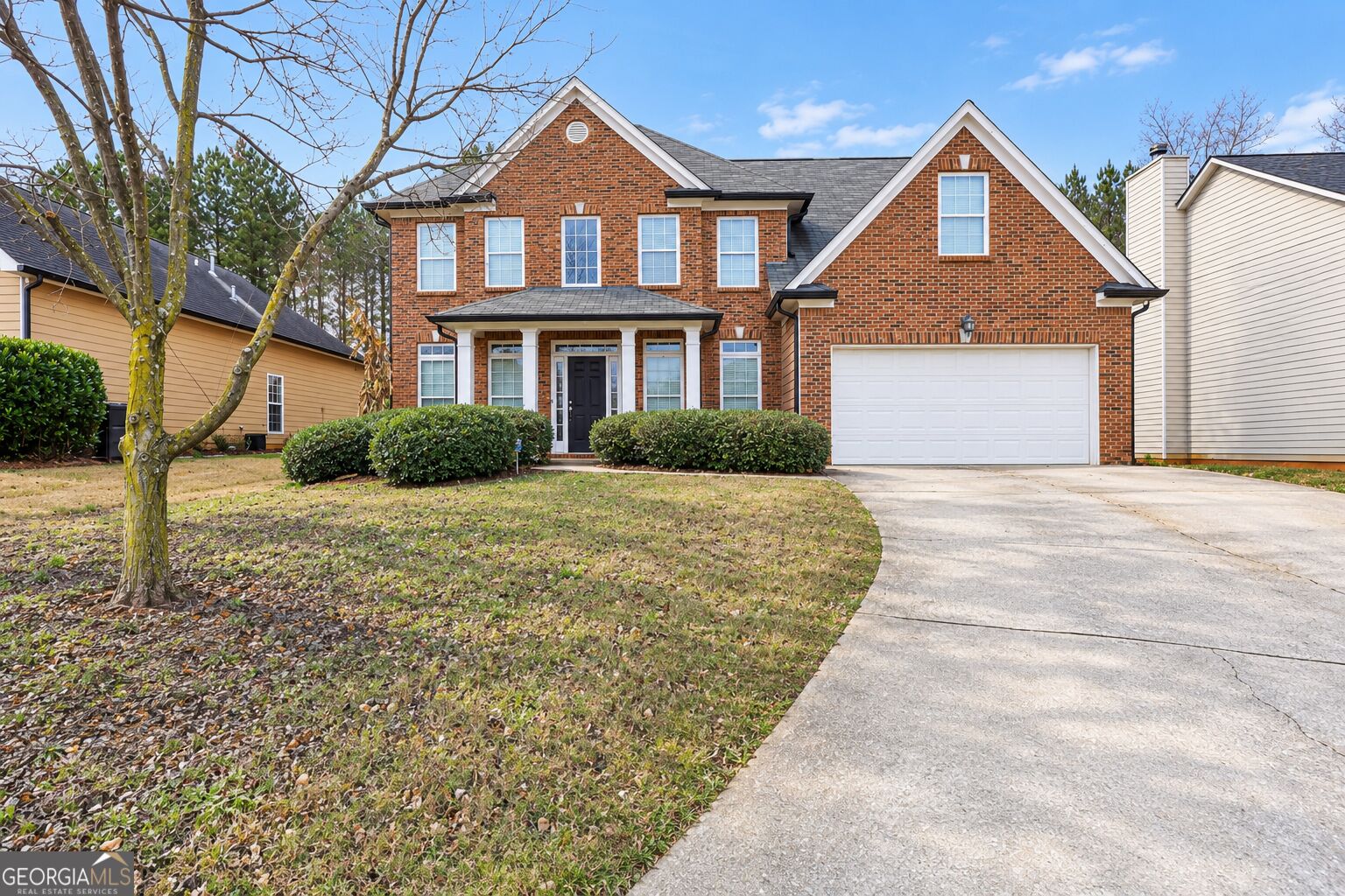 Two-story brick house with a white garage, broad driveway, and leafless tree in front.