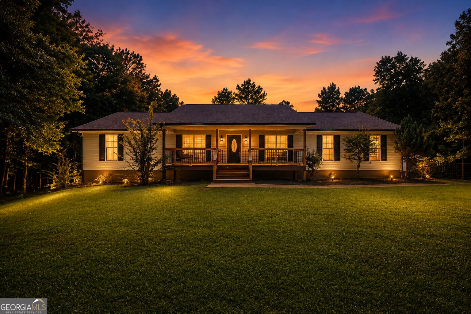 Single-story home with a broad covered porch, glowing windows, and a wooded lawn at dusk.
