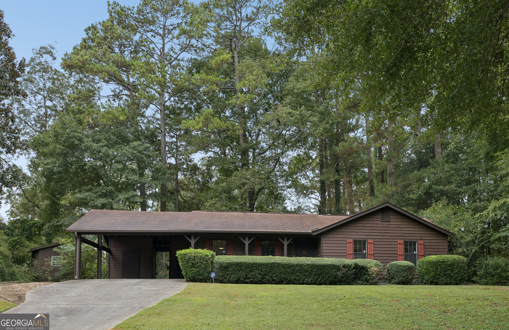 Brown single-story home with red shutters, a covered front porch, and tall trees behind it.