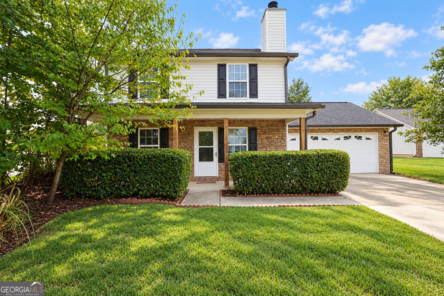 Two-story white and stone house with an attached garage, clipped hedges, and a broad front lawn.