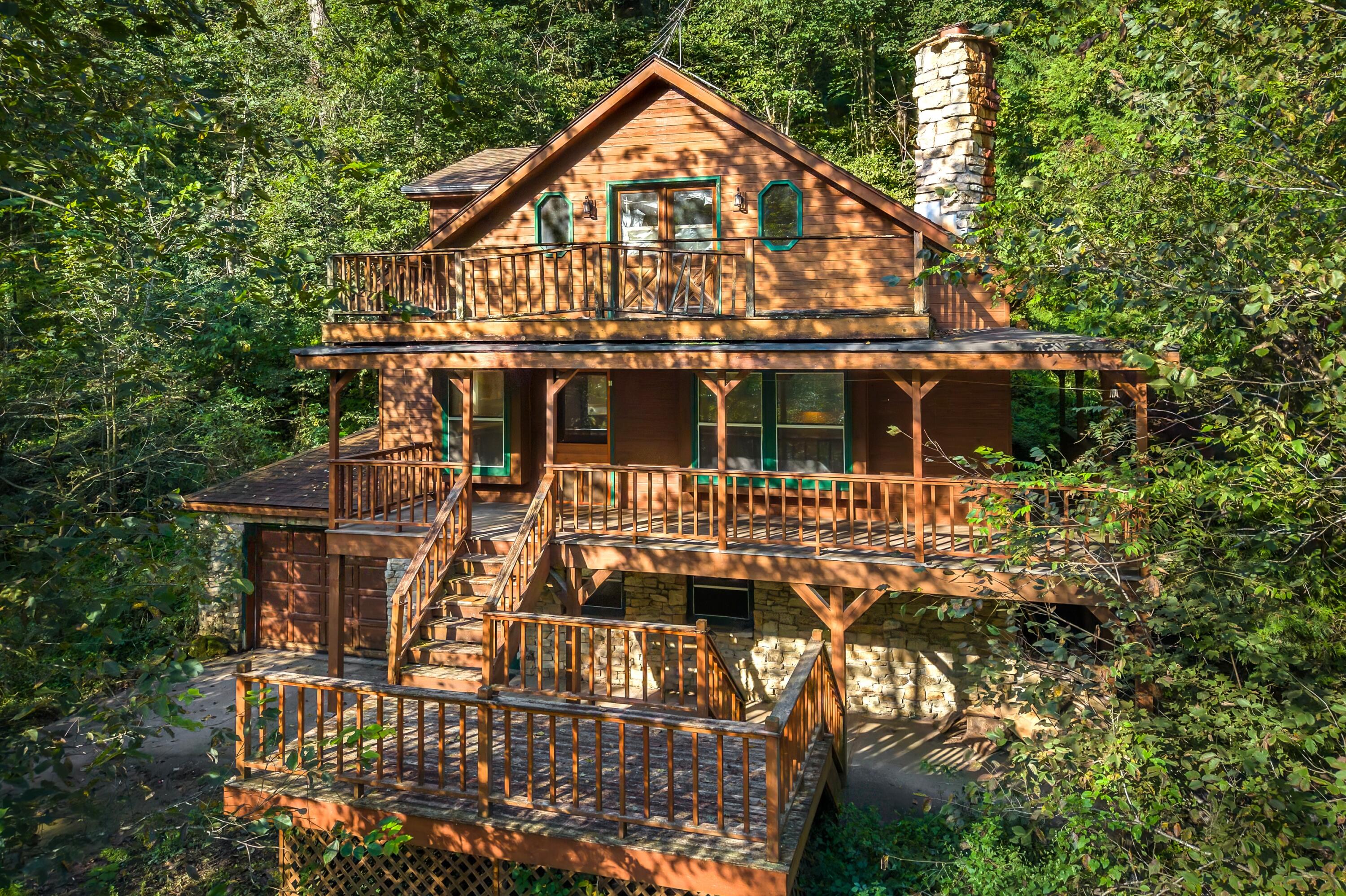 Wooded multi-level cabin with expansive wooden decks, stairways, stone foundation, and chimney.