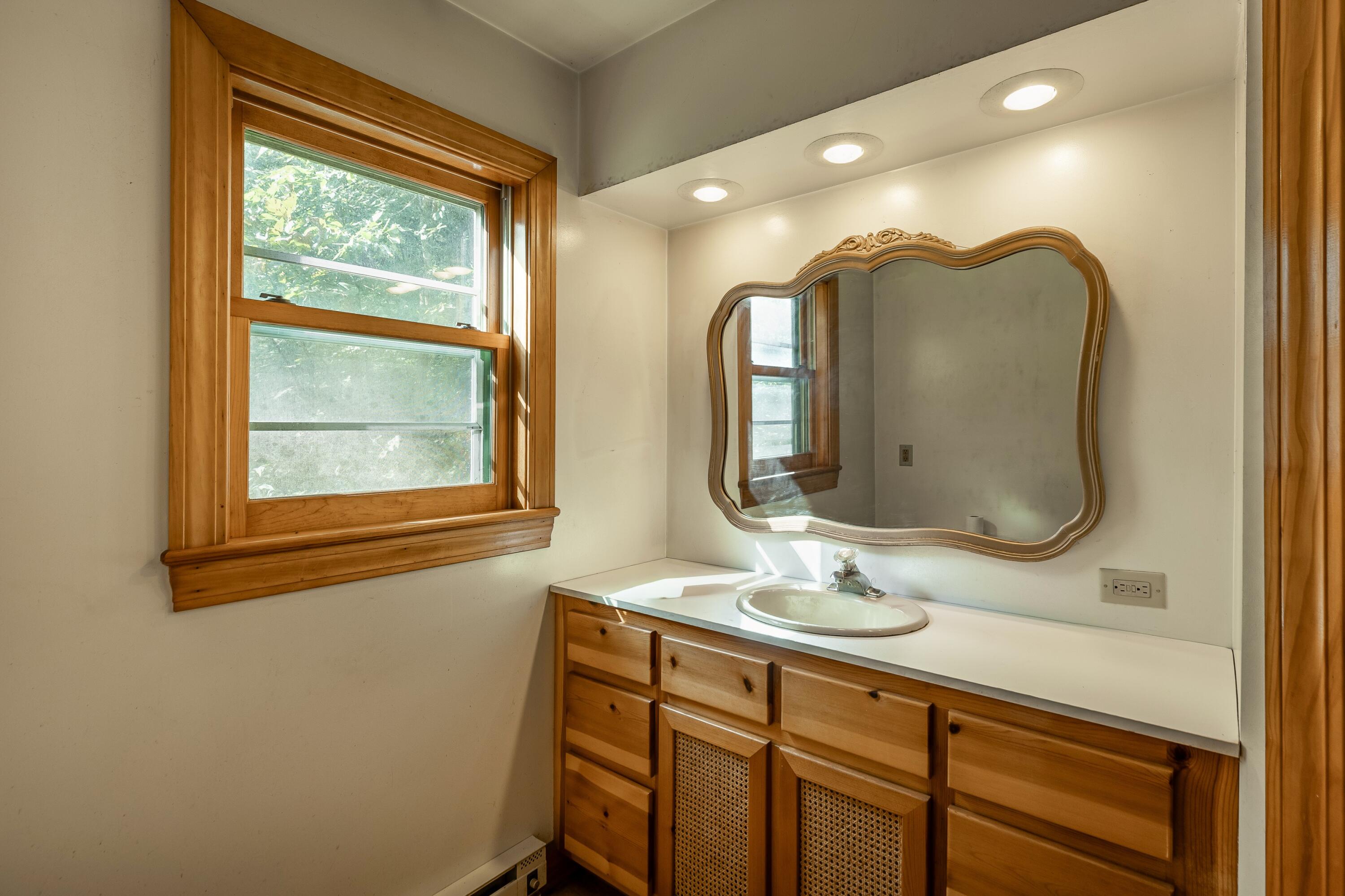 Bathroom vanity with ornate mirror, warm wood cabinets, and a wood-trimmed window.