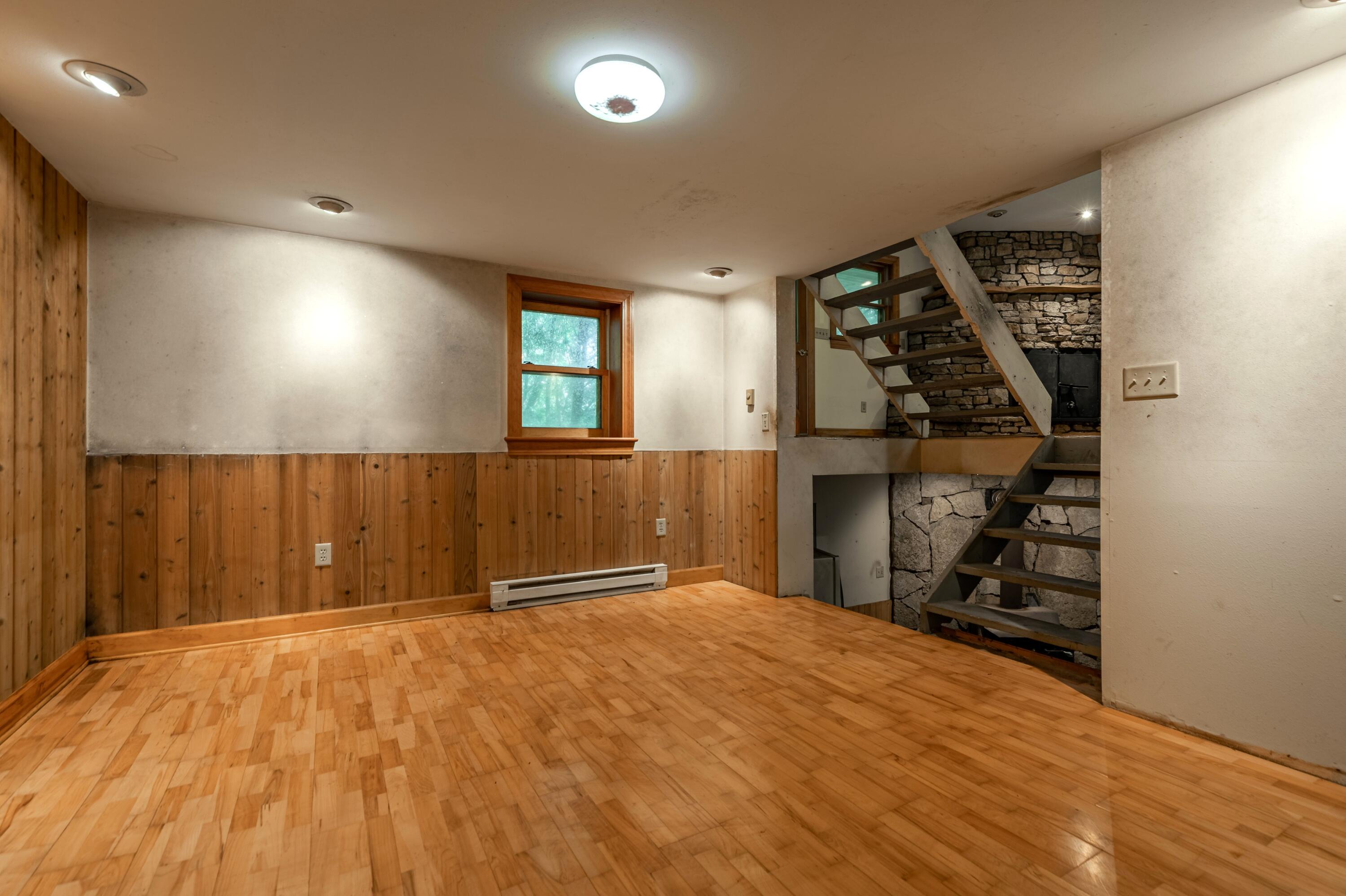 Empty room with hardwood floors, wood wainscoting, and an open staircase against a stone wall.