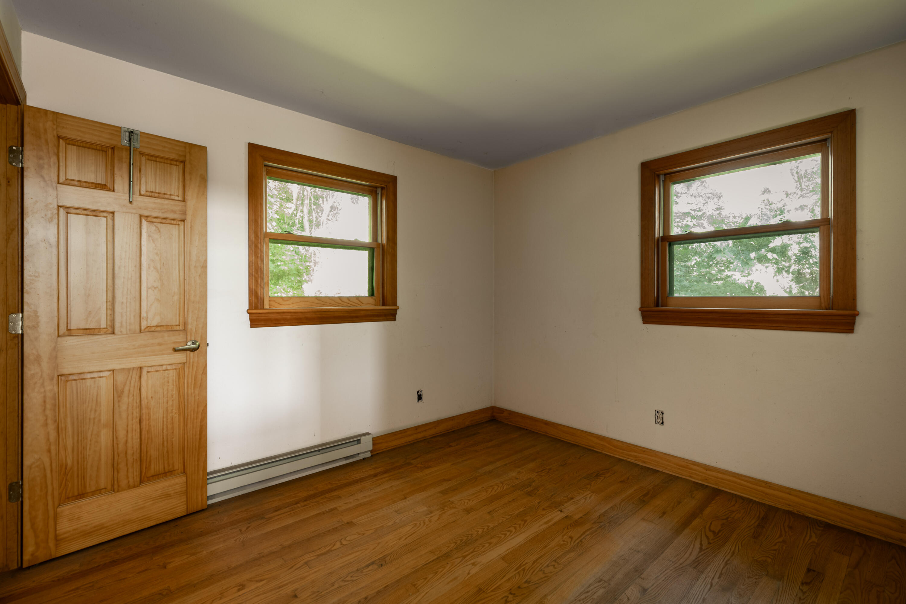 Empty room with hardwood floors, a paneled wooden door, and two wood-trimmed windows.