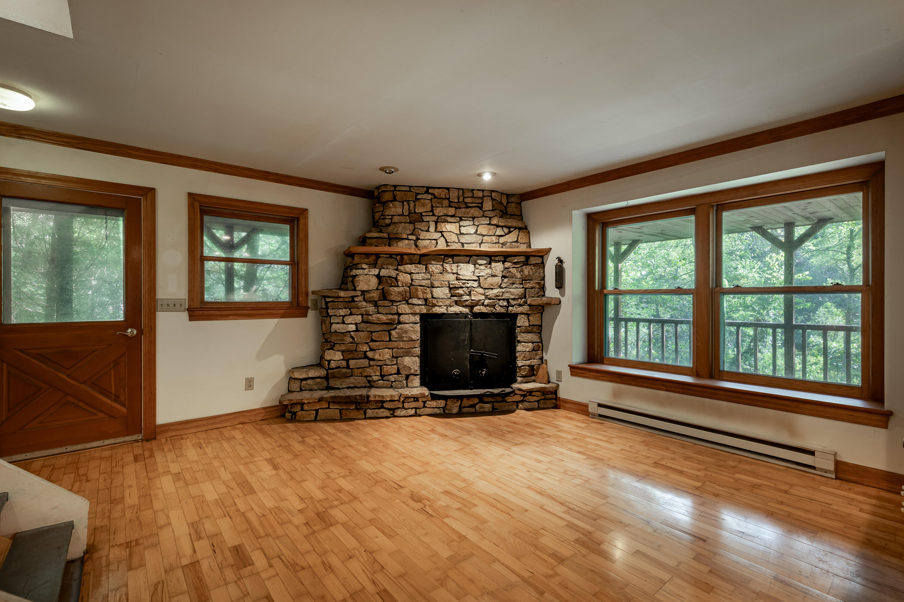 Empty living room with a broad stone fireplace, hardwood floors, and large windows overlooking trees.
