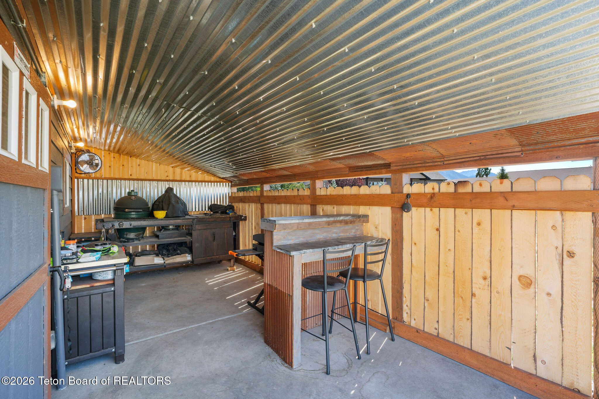 Covered patio with a grill and wooden bar seating beneath a corrugated metal roof.