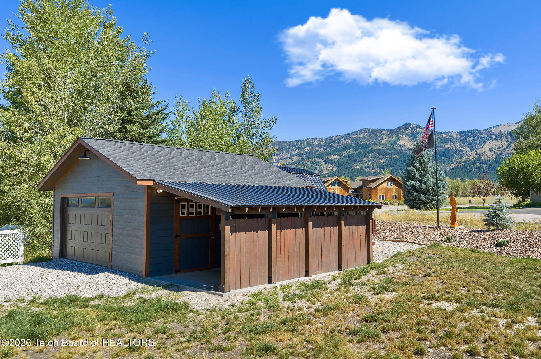 Detached garage with a long covered storage area, backed by trees and forested mountains.