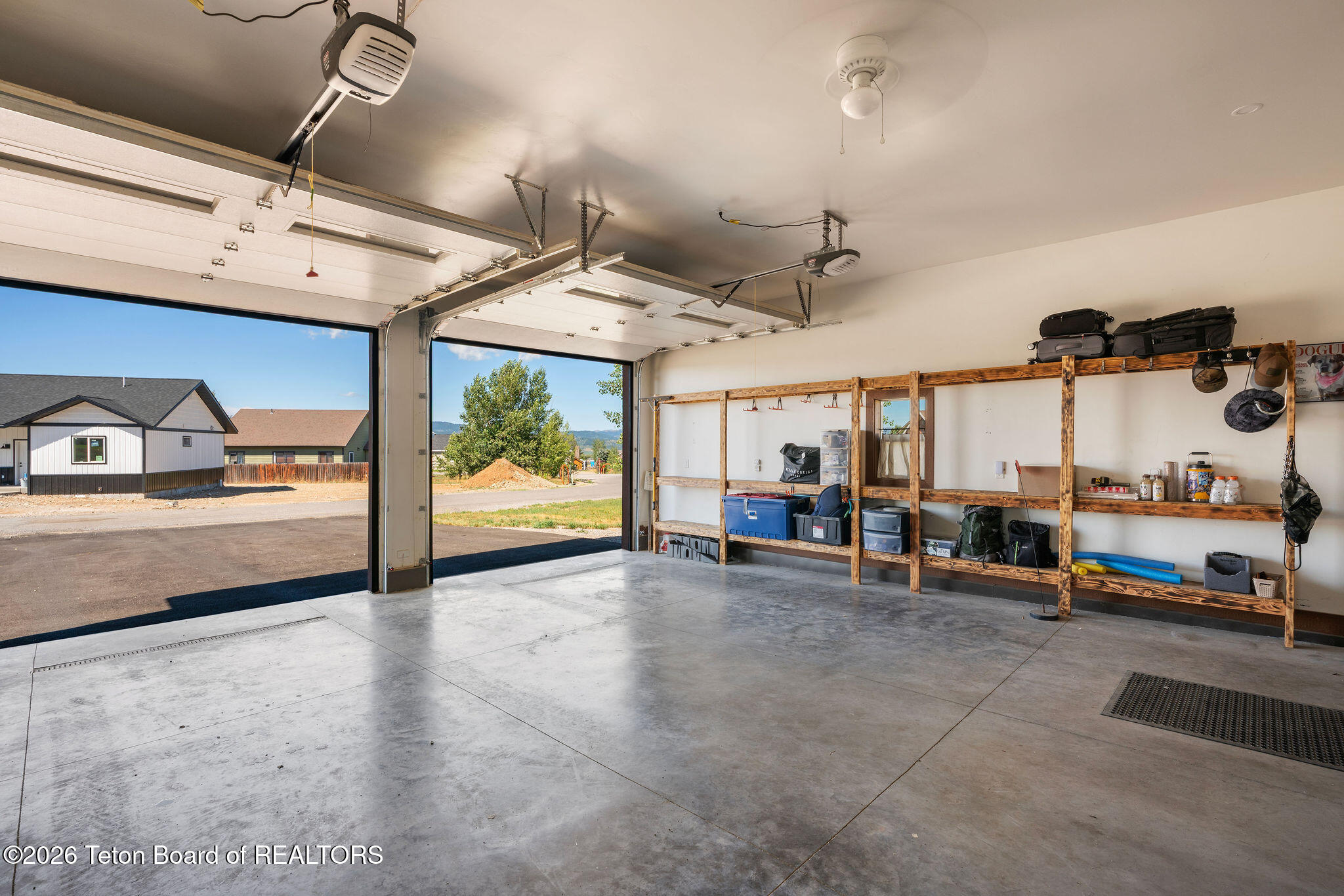 Two-car garage with open overhead doors, concrete floor, and long wooden storage shelves.