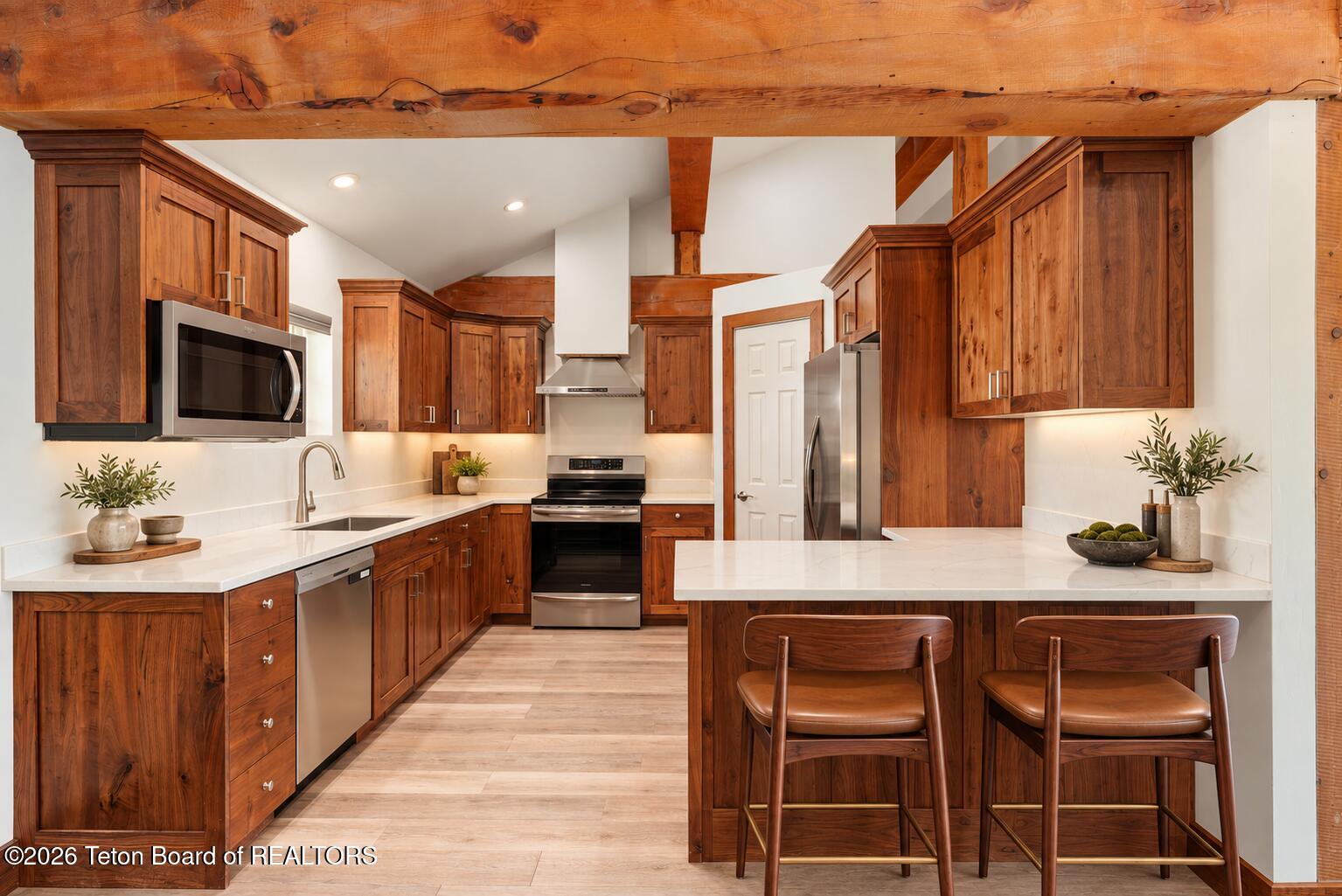 Kitchen with knotty wood cabinets, exposed beams, stainless steel appliances, and an island with two stools.