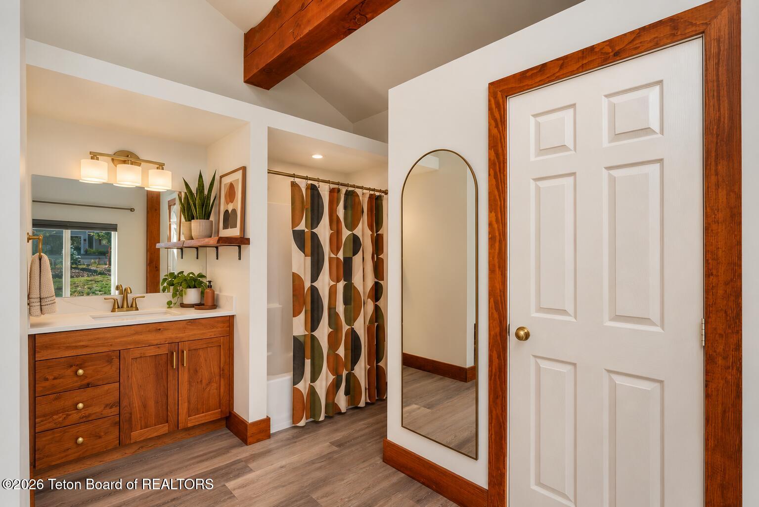 Bathroom with a warm wood vanity, patterned shower curtain, and arched full-length mirror.