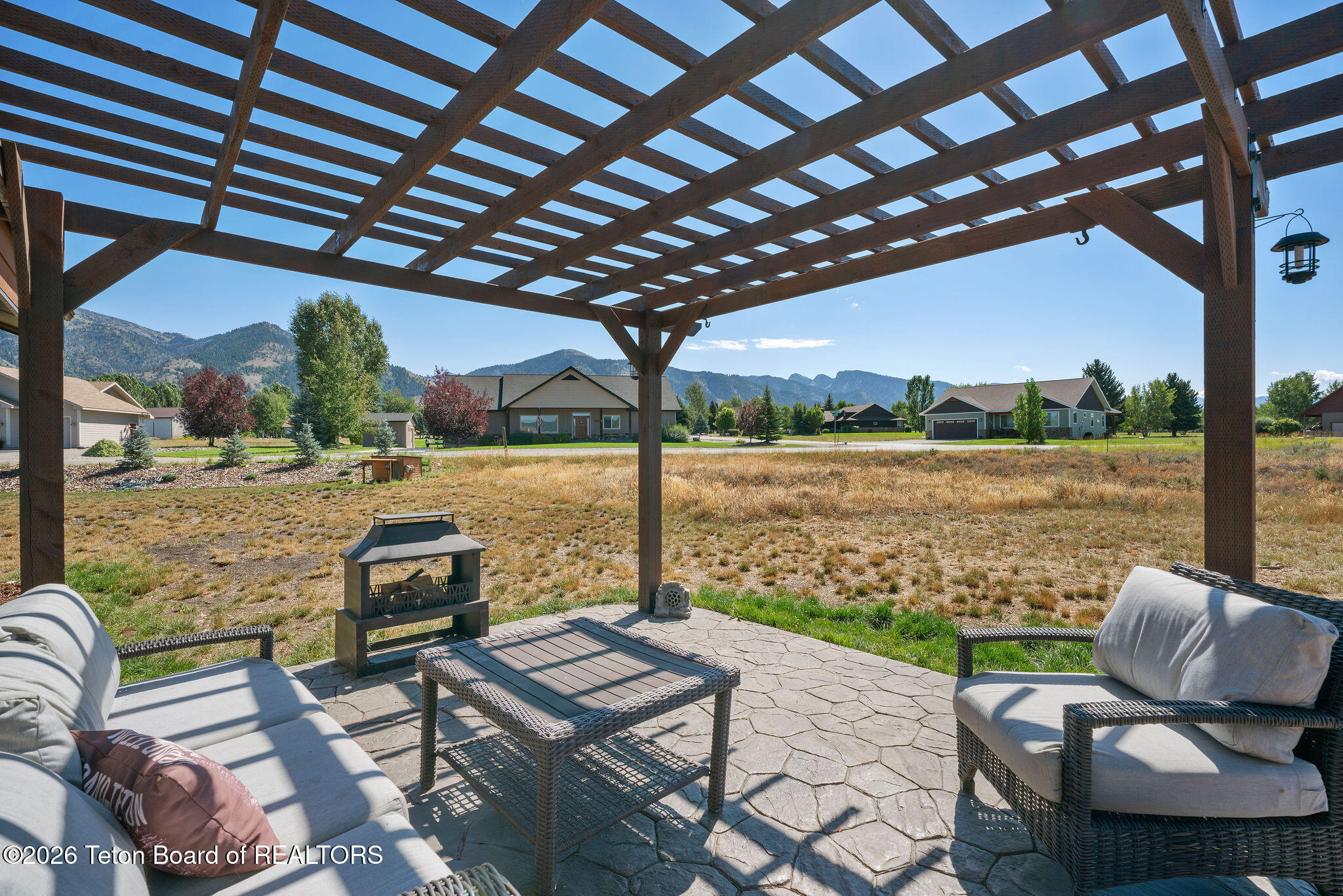 Furnished stone patio beneath a wooden pergola, overlooking a grassy yard and mountain ridges.