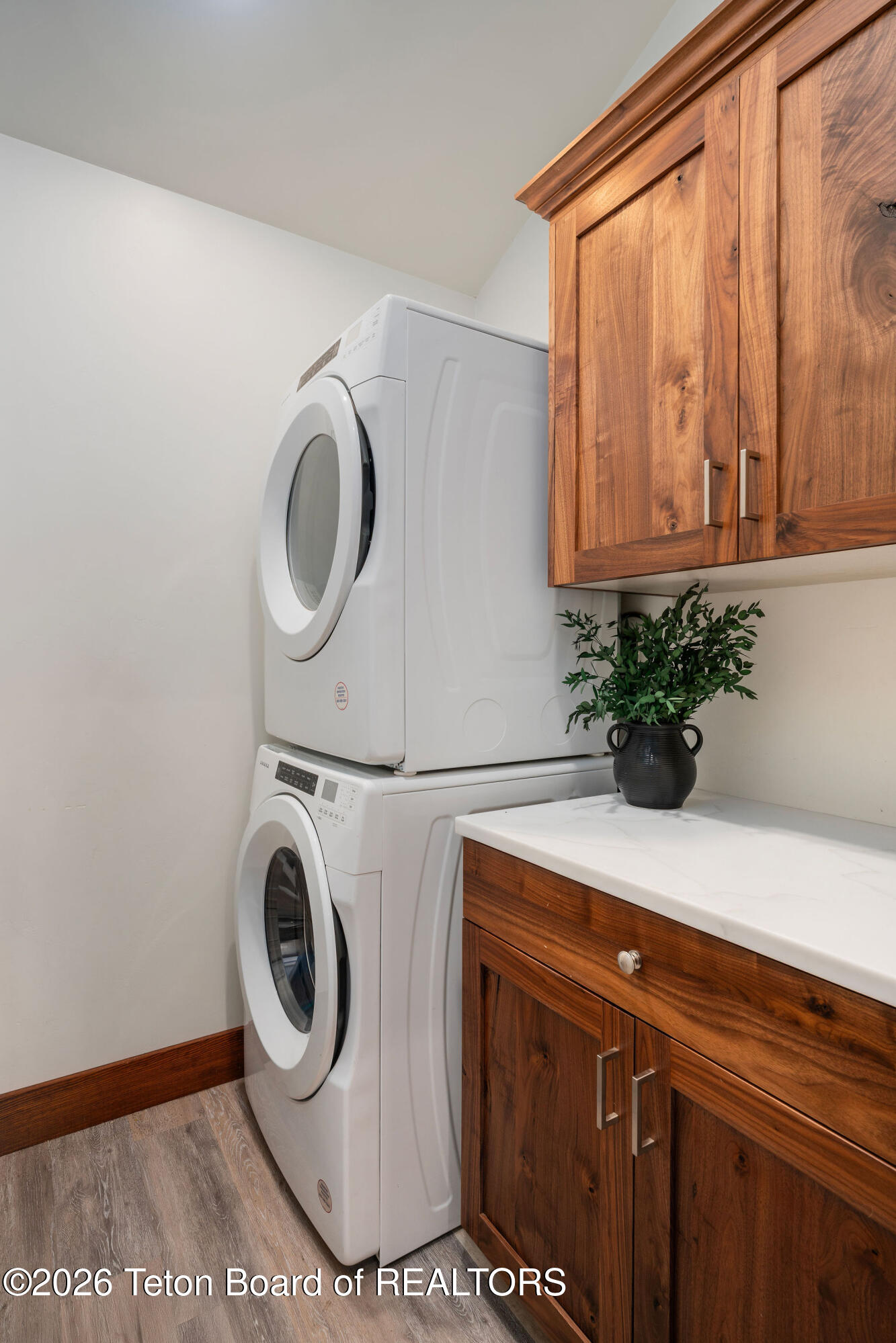 Laundry room with stacked front-loading washer and dryer beside warm wood cabinetry and a white countertop.