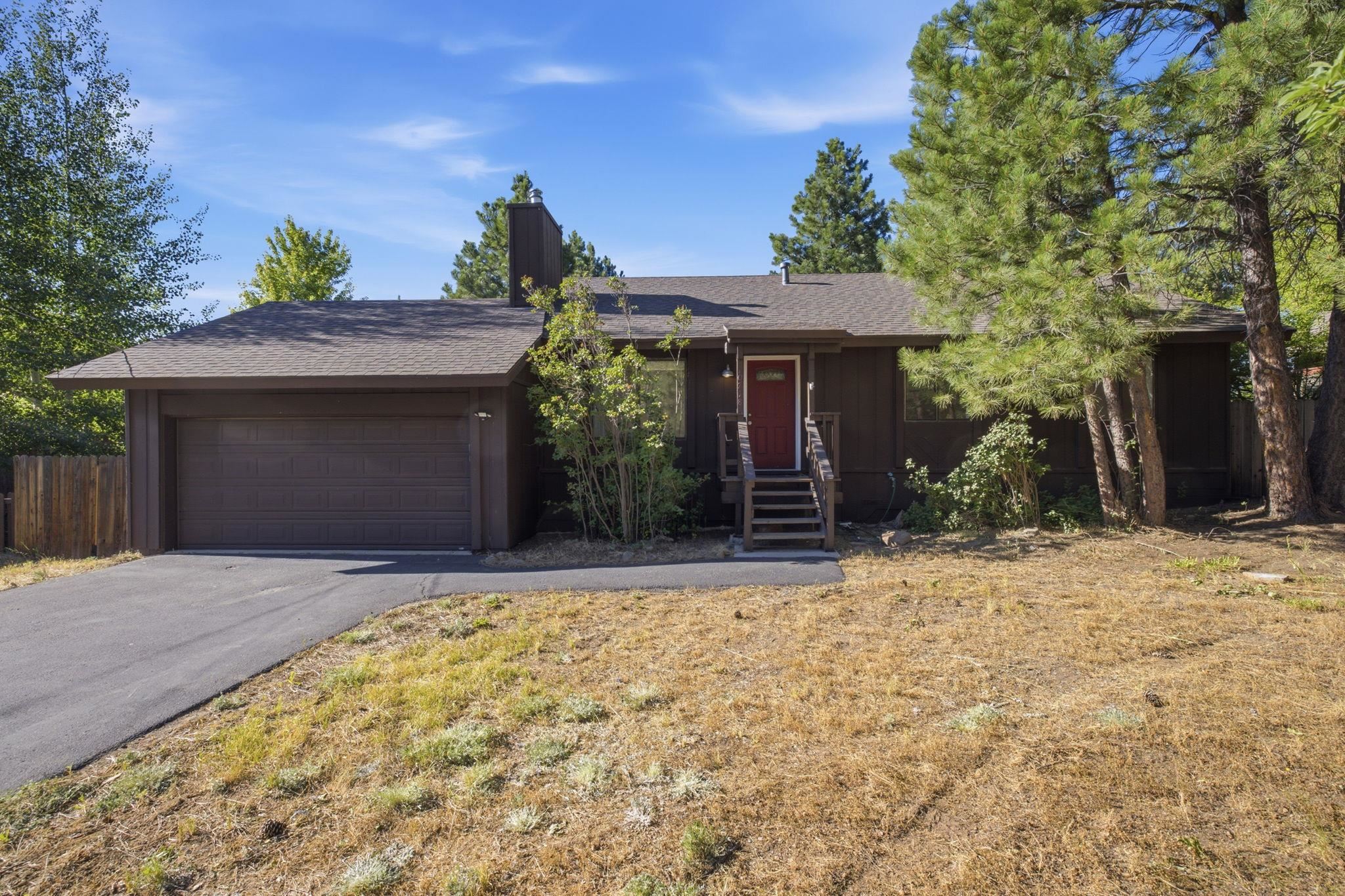 Dark brown single-story home with a two-car garage, red front door, and pine trees.