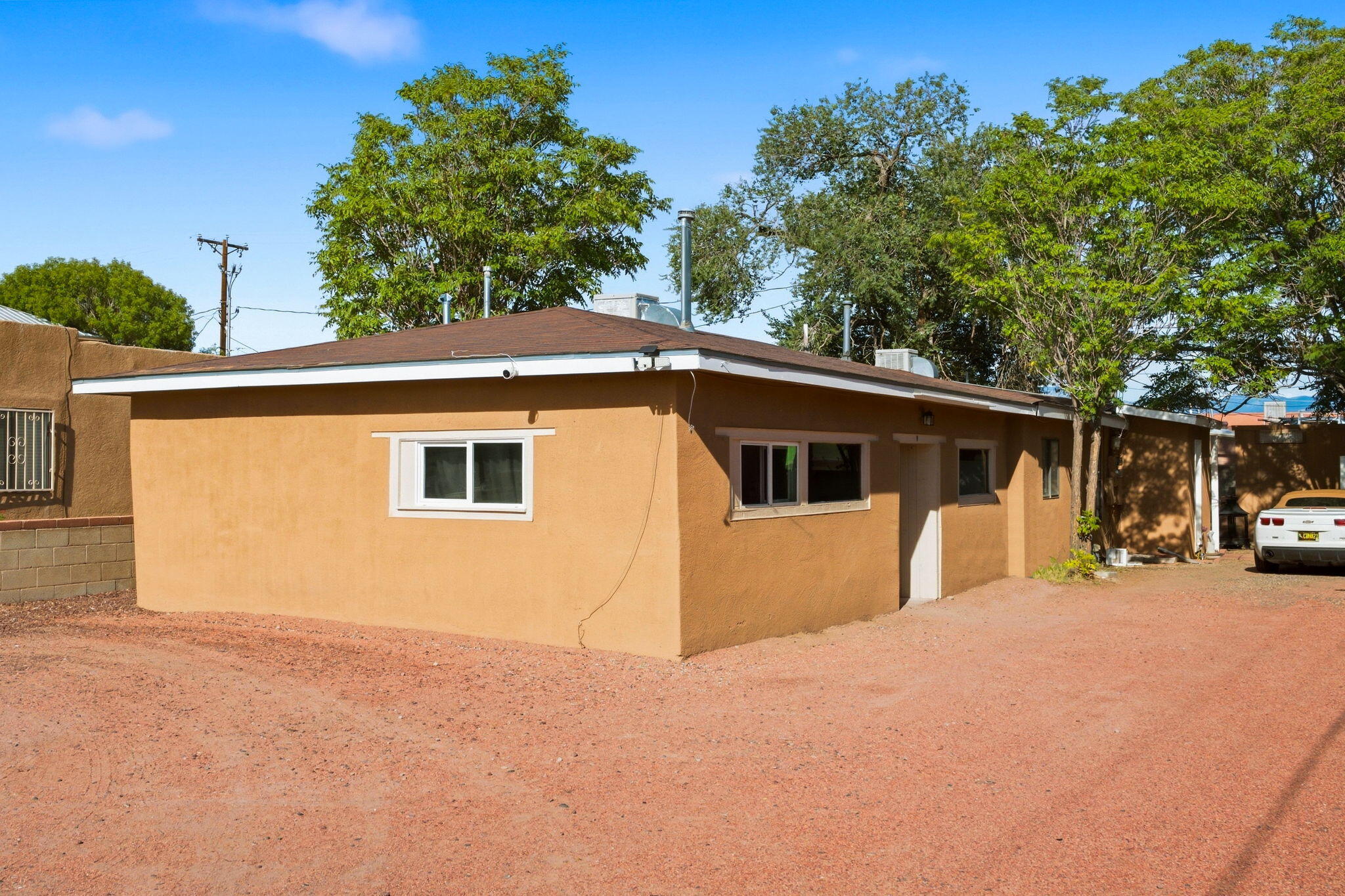 Tan stucco single-story home with a gravel yard, mature trees, and a white car.