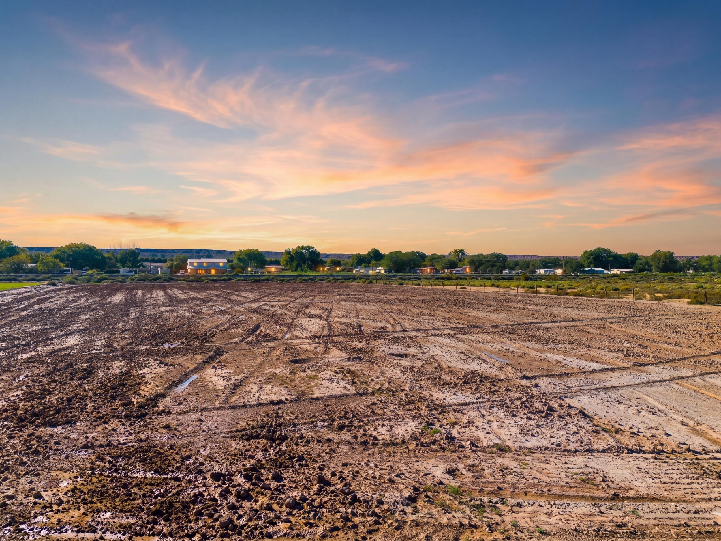 Vacant graded lot beneath a pink and orange sunset, with homes and trees in the distance.