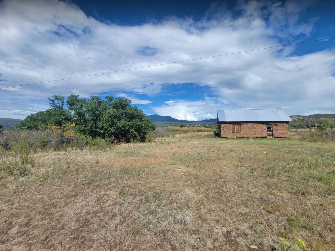 Adobe outbuilding with a metal roof in an open, dry field beneath distant mountains.
