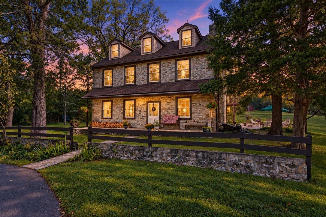 Stone farmhouse exterior with glowing windows and a fenced lawn at dusk.
