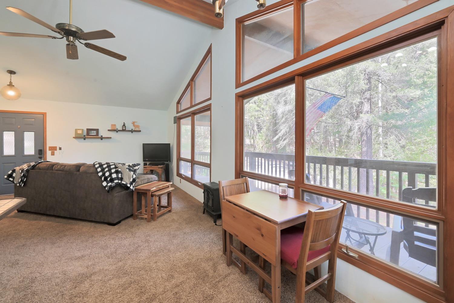 Living room with vaulted ceiling, wood-framed windows, and dining table overlooking a wooded deck.