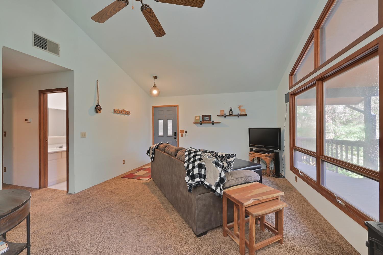 Living room with vaulted ceiling, brown sectional sofa, and large wood-trimmed windows overlooking a deck.