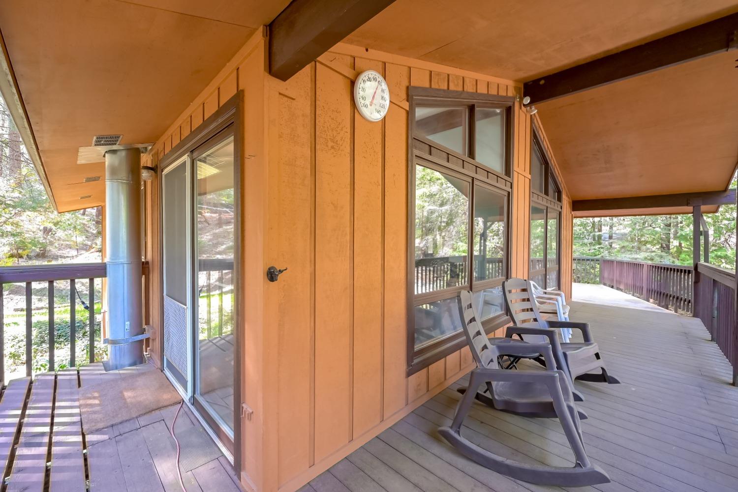 Covered wooden deck with sliding glass doors, large windows, and chairs overlooking trees.