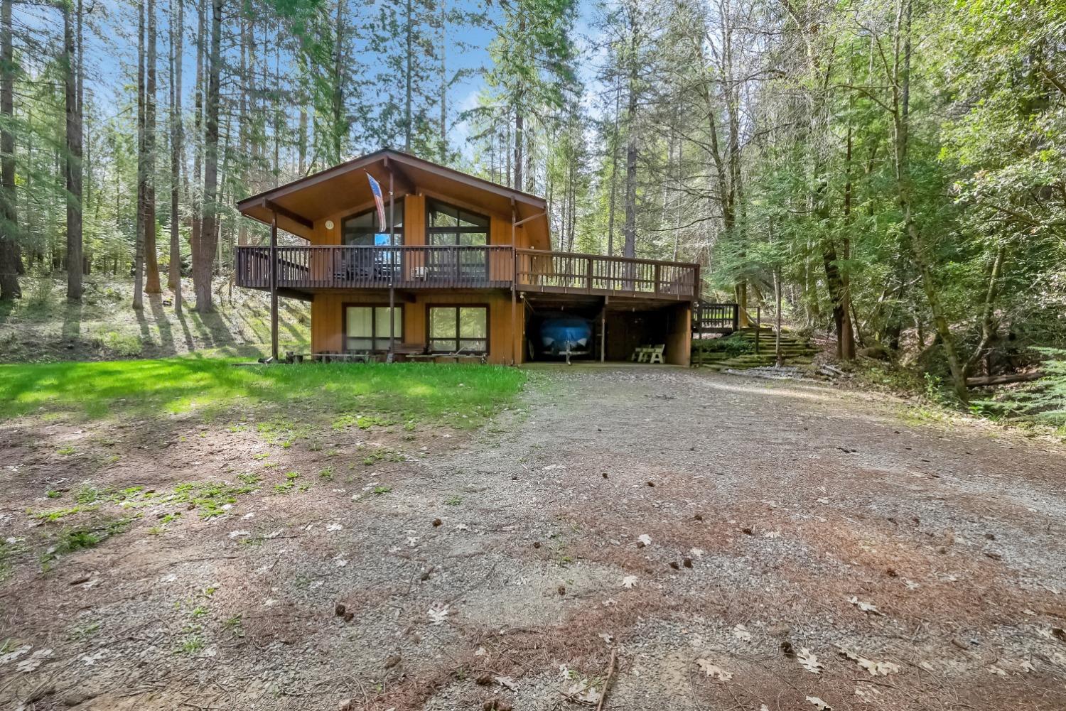 Wooded cabin with a wide raised deck, gravel driveway, and boat stored underneath.