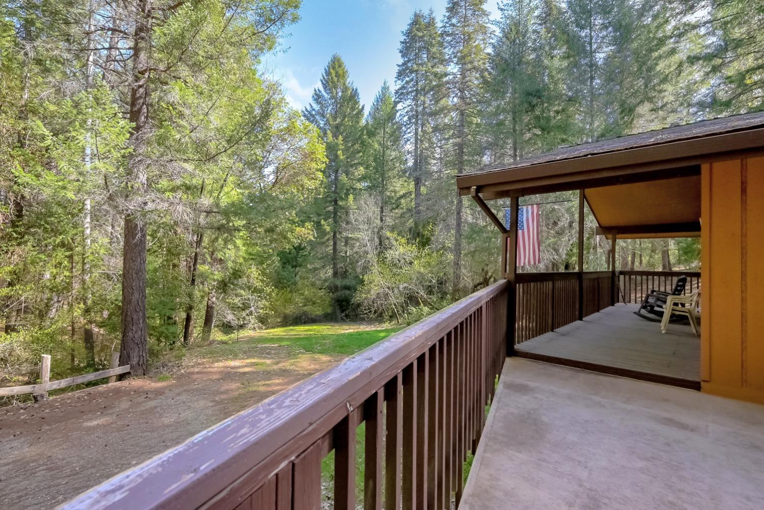 Wooden deck overlooking a forested clearing, with a covered porch, outdoor chairs, and an American flag.