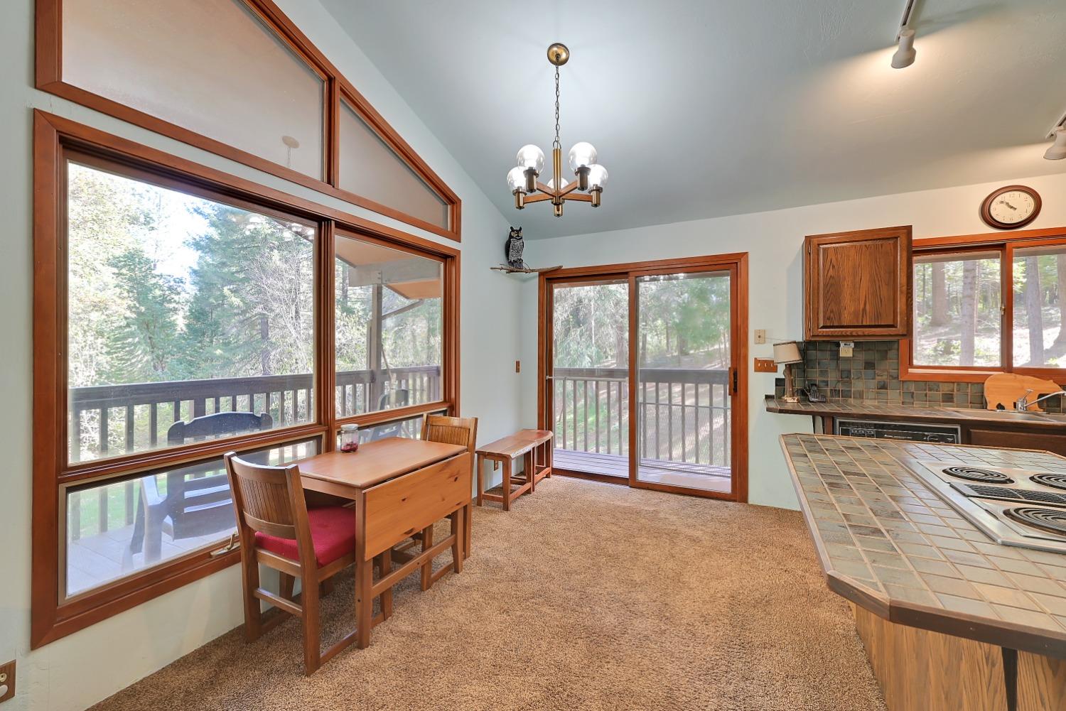 Dining area with a wood table beside large windows overlooking trees and a deck.