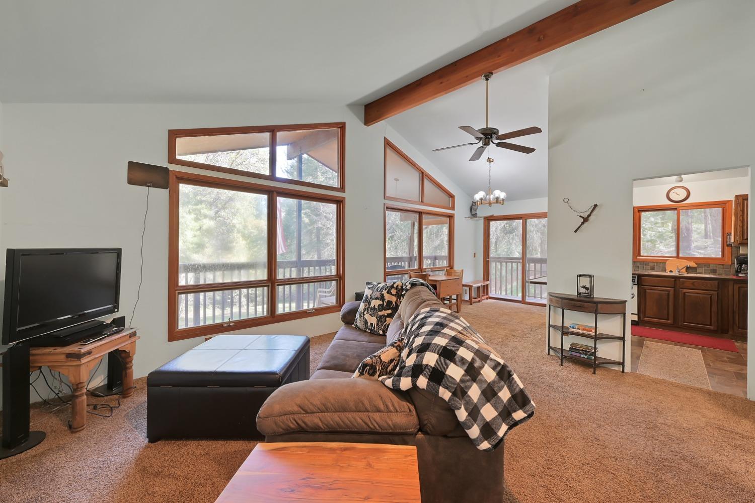 Living room with vaulted ceiling, exposed wood beam, and large windows overlooking trees.