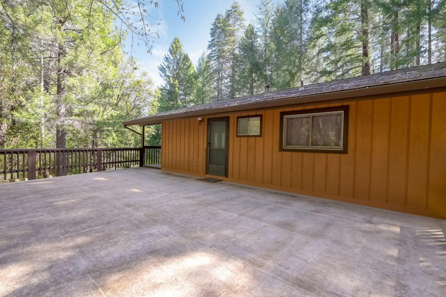 Concrete patio beside an orange-brown wood-sided home, with a deck railing and tall trees beyond.