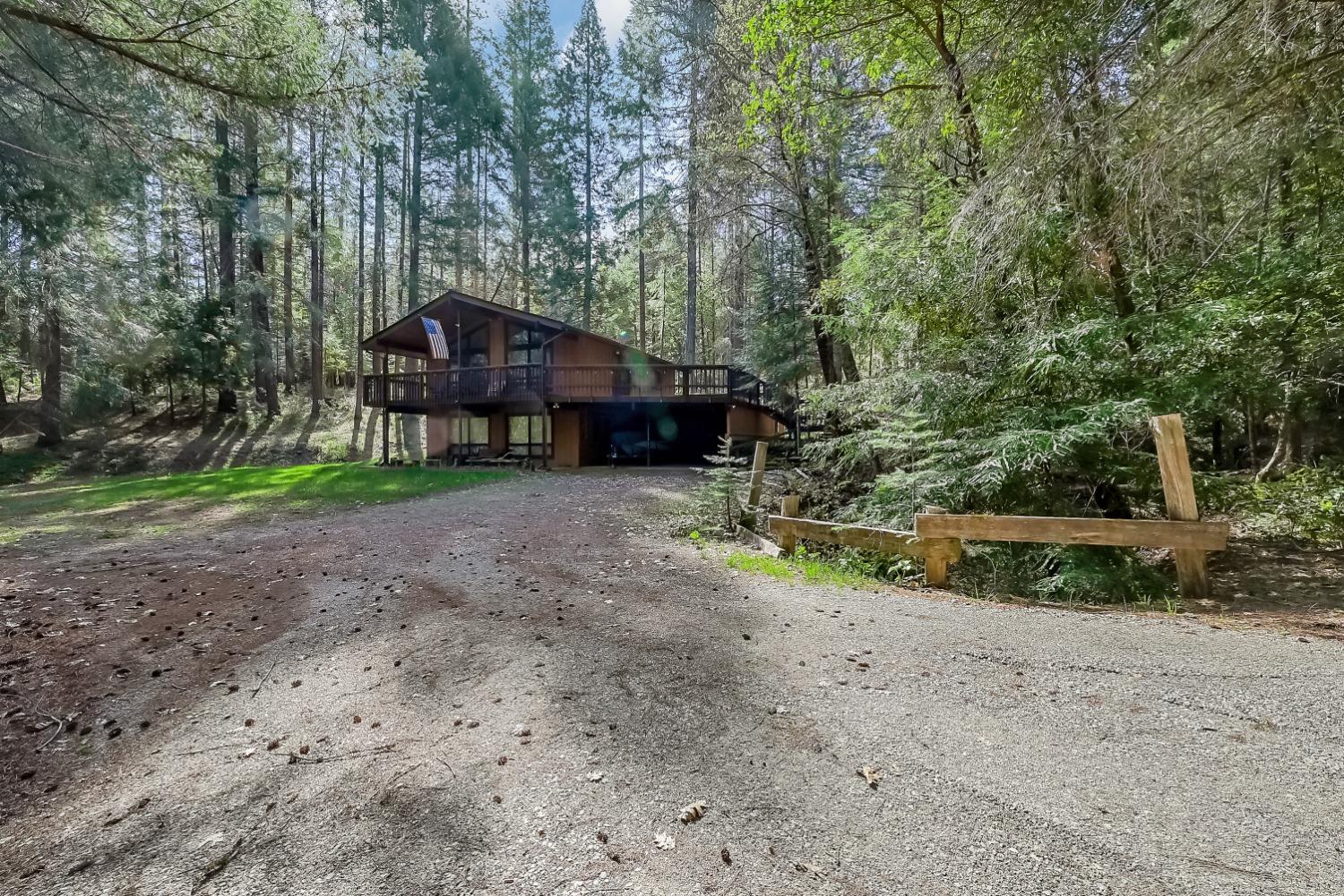 Wood-sided house with a raised deck nestled among tall trees at the end of a gravel driveway.