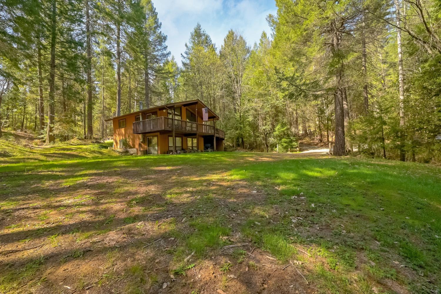 Two-story brown home with a raised deck in a sunny, wooded clearing.