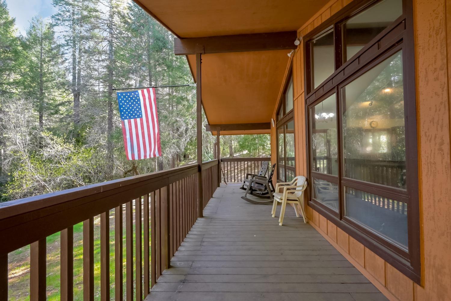 Covered wooden porch with brown railings, large windows, chairs, and an American flag among the trees.