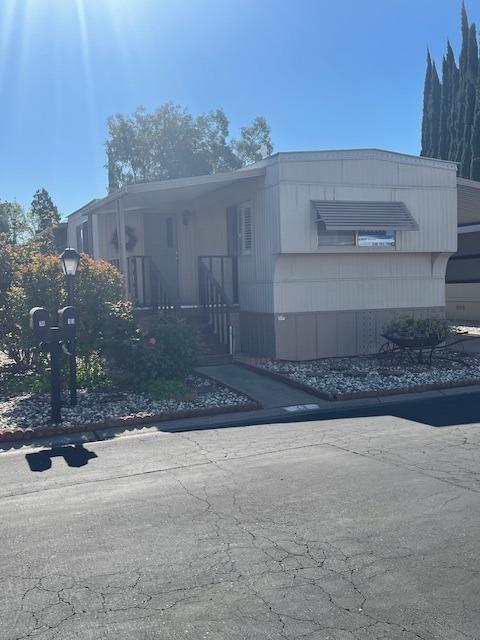 Light-colored manufactured home with a covered entry, window awning, and landscaped rock beds beside the street.
