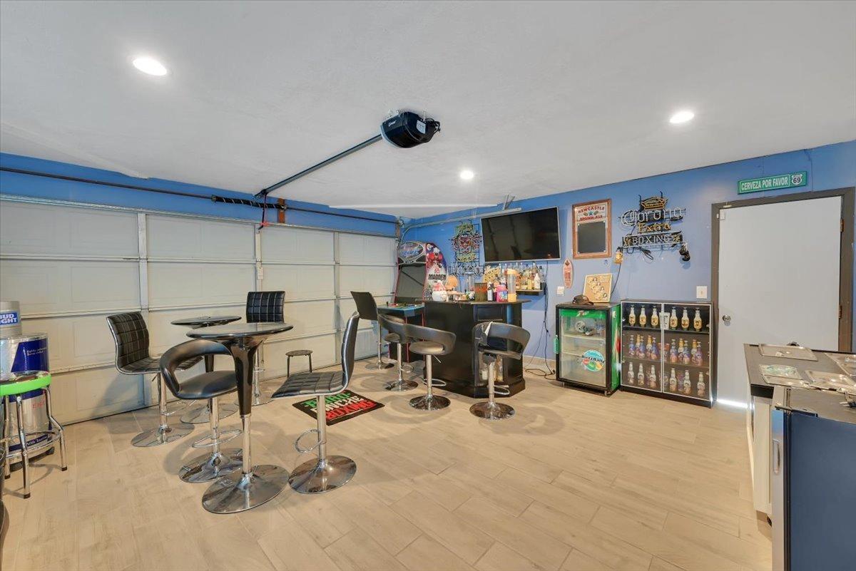 Garage bar with a counter, beverage refrigerators, television, stools, and closed overhead garage door.