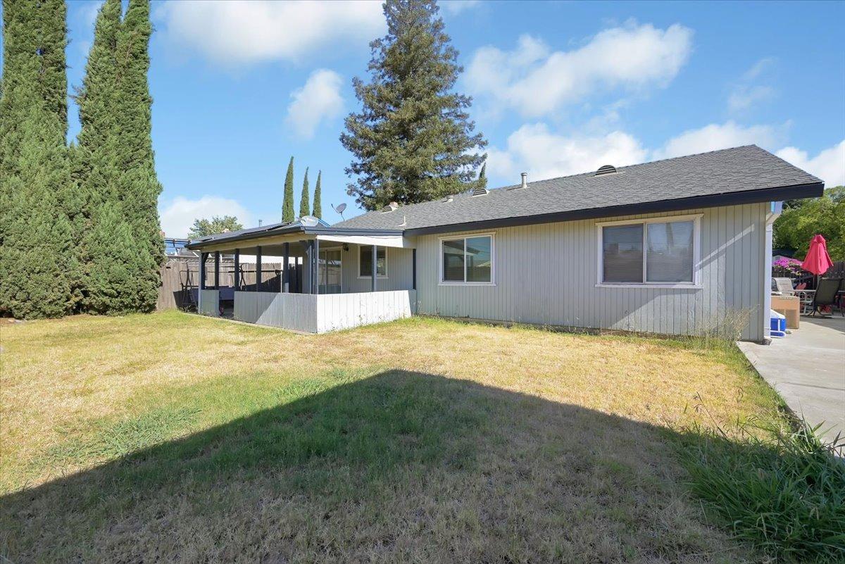 Single-story gray house with a covered patio, dry backyard lawn, and tall evergreen trees.