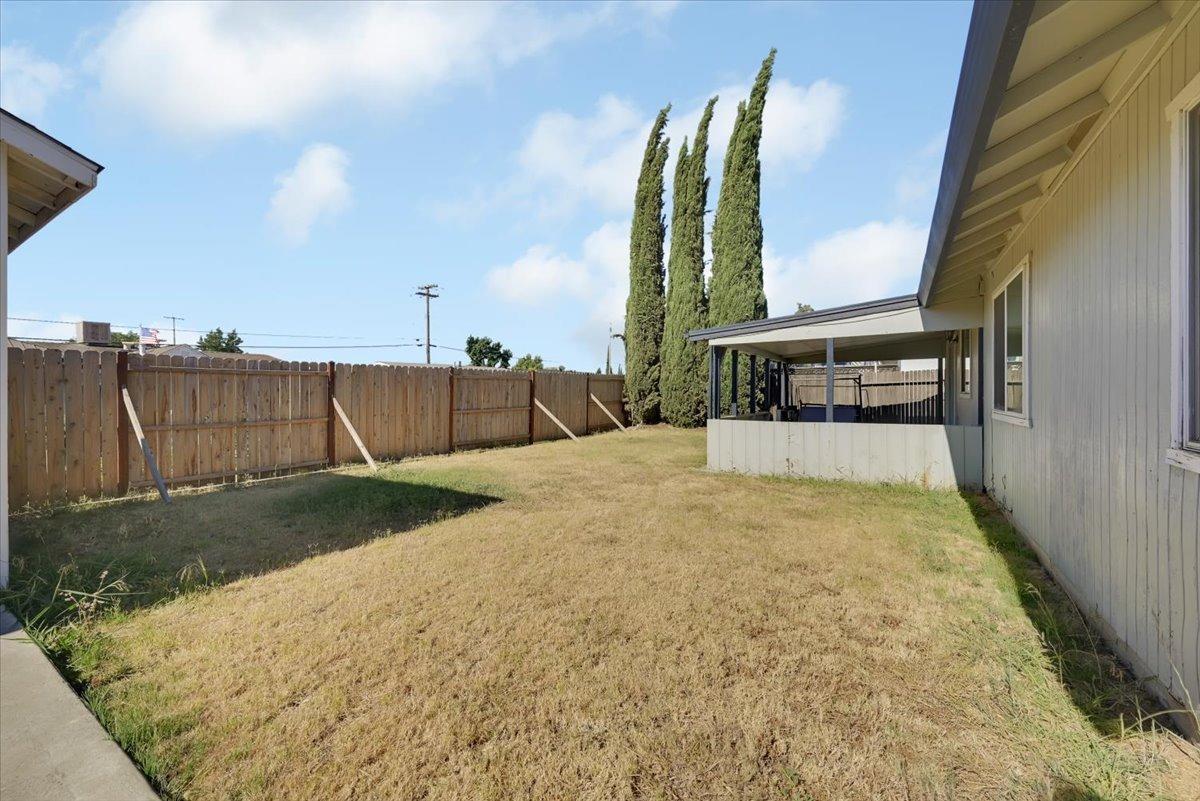 Fenced backyard with a covered patio, dry lawn, and tall cypress trees.