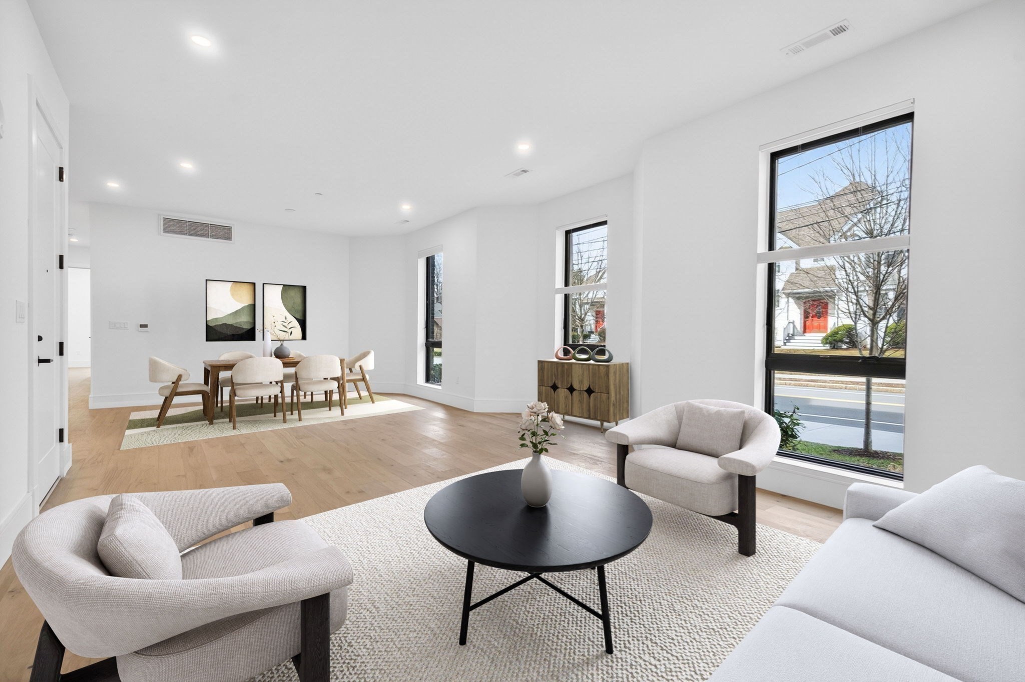 Open-plan living and dining room with neutral seating, light wood floors, and large street-facing windows.
